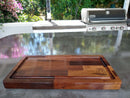 The Beast BBQ Board® 20" x 10" Cutting Board by BBQ Boards® sits on a sleek outdoor countertop, with a gas grill and greenery creating the perfect cookout setting.