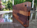 The Beast BBQ Board® 20" x 10" Cutting Board by BBQ Boards® stands upright on a reflective outdoor table, its varied wood tones framed by lush greenery and colorful flowers in the background.
