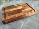An 18" x 12" Beastie BBQ Board® cutting board by BBQ Boards®, featuring a juice groove, mixed wood tones, and a “BBQ” logo with chef’s hat and utensils in one corner, rests on a smooth gray surface.