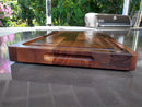 The BBQ Boards® Beastie BBQ Board® 18" x 12" Cutting Board with a handle slot and juice groove sits on a gray outdoor countertop, with green foliage and a barbecue grill in the background.