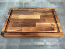 The Beastie BBQ Board® 18" x 12" Cutting Board by BBQ Boards® features a juice groove, mixed walnut tones, and an engraved rhino logo, shown on a grey concrete surface.