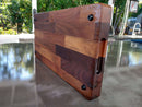 The Beastie BBQ Board® 18" x 12" Cutting Board by BBQ Boards® stands upright outdoors on a reflective surface. It features a patchwork design, juice groove, rounded edges, dark screws, and a built-in hand grip with trees in the background.