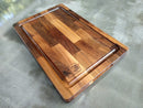 The Beastie BBQ Board® 18" x 12" Cutting Board by BBQ Boards® features a patchwork pattern of wood tones, a juice groove, and the BBQ Boards® logo engraved in one corner. It sits on a smooth gray surface.