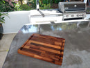 The Beastliest BBQ Board® 21" x 17" Cutting Board by BBQ Boards® rests on a shiny metal countertop in an outdoor kitchen, with a barbecue grill and lush greenery visible in the background.