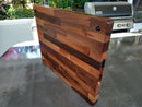 The BBQ Boards® Beastliest BBQ Board® 21" x 17" Cutting Board stands upright on a metallic outdoor countertop, with a barbecue grill and green plants in the background.