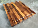 The Beastliest BBQ Board® 21" x 17" cutting board from BBQ Boards®, featuring alternating light and dark wood strips, a groove edge, and a "BBQ" logo, is shown on a smooth gray countertop.
