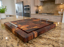 A BBQ Boards® Beefy BBQ Board® 18" x 15" cutting board with metal handles rests on a granite countertop in a modern kitchen with light wood cabinets, stainless steel appliances, and a potted plant in the background.