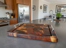 The Beefy BBQ Board® 18" x 15" Cutting Board from BBQ Boards® with metal handles sits on a kitchen island, surrounded by stainless steel appliances, wooden cabinets, and framed pictures in the background.