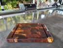 The Beefy BBQ Board® 18" x 15" Cutting Board by BBQ Boards®, featuring metal handles, sits on an outdoor countertop near a gas grill with greenery in the background.