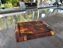 The Beefy BBQ Board® 18" x 15" Cutting Board by BBQ Boards® with metal handles rests on a sleek outdoor kitchen counter, while a grill and sink are visible in the background amid lush greenery.