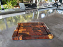 The BBQ Boards® Beefy BBQ Board® 18" x 15" Cutting Board with metal handles rests on a sleek outdoor countertop, serving as both a prep and serving tray near a stainless steel gas grill and vibrant greenery.