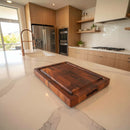 The Best BBQ Board® 16 x 12 Cutting Board by BBQ Boards® rests on a white marble kitchen island in a modern space with wood cabinets, stainless steel appliances, and a gas stove in the background.