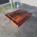 The Best BBQ Board® 16 x 12 Cutting Board by BBQ Boards®, featuring a herringbone pattern and juice groove, adds warmth with its rich brown tones on a reflective gray countertop, contrasting the smooth metallic surface beneath.