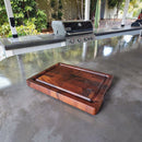 The Best BBQ Board® 16 x 12 Cutting Board by BBQ Boards® sits on a shiny outdoor countertop, with a barbecue grill and greenery in the background.