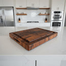 The Best BBQ Board® 16 x 12 Cutting Board by BBQ Boards®, featuring a checkered pattern, rests on a white marble kitchen island with modern white cabinets, stainless steel appliances, and wooden shelves in the background.