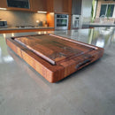 The BBQ Boards® Best BBQ Board® 16 x 12 Cutting Board, featuring curved edges and a juice groove, rests on a shiny kitchen island with modern appliances and cabinets in the background.