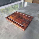 The BBQ Boards® Best BBQ Board® 16 x 12 Cutting Board features a durable patchwork wood grain, edge groove, engraved logo, and rectangular shape—perfect as your go-to BBQ cutting board on any shiny grey surface.