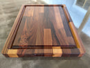 An 18 x 15-inch reversible Big BBQ Board® by BBQ Boards® with grooved edges and alternating light and dark wood tones rests on a reflective countertop, featuring the brand’s logo in one corner.