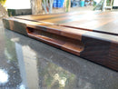 Close-up of the BBQ Boards® Bigger BBQ Board® 24" x 24" Reversible Cutting Board, featuring a grooved handle and juice groove, placed on a wet outdoor surface with blurred trees and containers in the background.