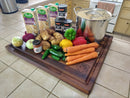 The BBQ Boards® Bigger BBQ Board® 24" x 24" Reversible Cutting Board on the kitchen counter holds carrots, potatoes, onions, bell peppers, jalapeños, canned coconut milk, boxed chicken broth, curry powder, and a large stock pot.