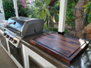 A BBQ Boards® Bigger BBQ Board® 24" x 24" Reversible Cutting Board rests on an outdoor kitchen counter near a stainless steel gas grill, surrounded by prickly pear cactus and blooming bougainvillea.