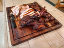Sliced smoked meat, whole cooked potatoes, and a knife rest on the BBQ Boards® Bigger BBQ Board® 24" x 24" reversible cutting board atop a tiled kitchen counter.