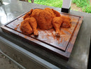 A whole spatchcocked chicken with red seasoning rests on the BBQ Boards® Bigger BBQ Board® 24" x 24" Reversible Cutting Board atop an outdoor kitchen counter, with greenery visible in the background.