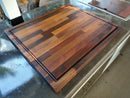 The Bigger BBQ Board® 24" x 24" Reversible Cutting Board by BBQ Boards® sits on a shiny outdoor countertop near a built-in grill. Its patterned wood in varied browns and juice groove makes it ideal for grilling enthusiasts.