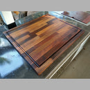 The BBQ Boards® Bigger BBQ Board® 24" x 24" Reversible Cutting Board, featuring a rich, patterned grain, rests on a shiny outdoor countertop beside a built-in grill in a natural outdoor setting.