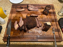 The BBQ Boards® Bigger BBQ Board® 24" x 24" Reversible Cutting Board displays sliced and chunked smoked brisket, with a knife on the left, a fork on the right, and mustard plus kitchen items in the background.