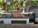 An outdoor kitchen features a sink, grill, leafy plants, and a decorative sign. On the counter is the BBQ Boards® Biggest BBQ Board® 36 x 24 Reversible Cutting Board, with a hanging plant above the area.