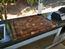 The Biggest BBQ Board® 36 x 24 Reversible Cutting Board by BBQ Boards® stands out on a sleek outdoor counter near a sink and grill, shaded partly by surrounding plants.