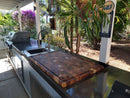 Outdoor kitchen features the BBQ Boards® Biggest BBQ Board® 36 x 24 Reversible Cutting Board, plus a sink and grill on a sleek counter. Surrounded by greenery, with a vintage Gulf sign hanging on the support post.