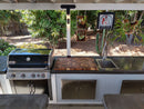 An outdoor kitchen features a grill, sink, metal cupboards, and the BBQ Boards® Biggest BBQ Board® 36 x 24 Reversible Cutting Board. A vintage "Beware of Lions" sign hangs above the sink, framed by lush greenery and a cactus in the background.