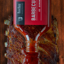 Bourbon Barrel Foods Barbecue Sauce with Bourbon and Sorghum is being poured over a rack of cooked ribs on a wooden surface, glazing the meat.