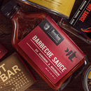 A close-up of Bourbon Barrel Foods Barbecue Sauce with Bourbon and Sorghum, featuring a red label, placed among other food products.