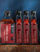 Four bottles of Bourbon Barrel Foods Barbecue Sauce with Bourbon and Sorghum—three tall, one square—sit on a wooden surface against a dark blue background. The labels display the Bourbon Barrel Foods brand.