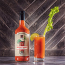 A bottle of Bourbon Barrel Foods Barrel Aged & Bourbon Smoked Bloody Mary Mix sits beside a Bloody Mary garnished with celery and lime, all set against a dark wooden background.