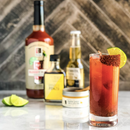A tall glass of reddish Bourbon Barrel Foods Barrel Aged & Bourbon Smoked Bloody Mary Mix, rimmed with chili powder and garnished with a lime wedge, sits on marble. Behind it are bottles, ingredient jars, and extra lime wedges nearby.
