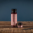 Two jars of Bourbon Barrel Foods Black Raspberry Jam, one tall and one small, sit on a rustic wooden surface against a plain blue background. Both jars feature pink labels, highlighting true Appalachian fruit flavor.