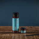 Two jars of Bourbon Barrel Foods Blueberry Bourbon & Cardamom Jam, one tall and one small with blue labels, sit on a wooden surface against a deep blue background—handcrafted in Kentucky for rich, authentic flavor.