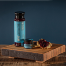 A tall bottle and small jar of Bourbon Barrel Foods Blueberry Bourbon & Cardamom Jam are displayed on a wooden board next to a biscuit topped with jam, all set against a dark blue background.