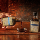 A hand pours Bourbon Barrel Foods Bluegrass Soy Sauce from a labeled glass bottle into a dish, with another identical bottle nearby on a rustic wooden surface, highlighting its rich umami flavor.