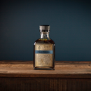 A glass bottle of Bourbon Barrel Foods Bluegrass Soy Sauce with a black cap sits on a rustic wooden surface against a dark blue background, its label highlighting the handcrafted details and rich umami flavor from Bourbon Barrel Foods.