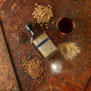 A bottle of Bourbon Barrel Foods Bluegrass Soy Sauce rests on a rustic surface with whole soybeans, wheat grains, a glass of soy sauce, salt, and ground ginger, showcasing its rich umami flavor and bourbon barrel aging.