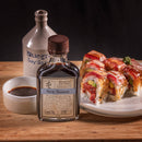 A bottle of Bourbon Barrel Foods Bluegrass Soy Sauce with a ceramic jug, a small dish of this soy sauce, and assorted sushi rolls on wood highlight the rich umami flavor of Bourbon Barrel Foods' signature product.