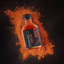 A bottle of Bourbon Barrel Foods Bourbon Barrel Aged Hot Sauce with a black label rests on a dark surface, surrounded by vibrant red chili powder. The label shows a chili pepper illustration and reads "HOT SAUCE.