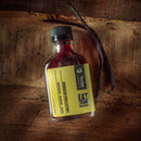 A square glass bottle of Bourbon Barrel Foods Bourbon Barrel Aged Madagascar Vanilla Extract with a gold label sits on a rustic wooden surface next to two vanilla beans.