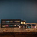 Bourbon Barrel Foods’ Bourbon Barrel Jams Gift Set, featuring five flavors like maple syrup, sorghum, bourbon barrel jams, and apple butter, is displayed in small jars on a rustic wooden surface against a dark blue background.