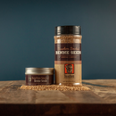 Two containers of Bourbon Barrel Foods Bourbon Smoked Benne Seeds—a small tin and a large shaker—rest on rustic wood, highlighted by scattered seeds against a blue background, showcasing this unique product from Bourbon Barrel Foods.